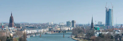 Ein Panoramablick auf eine Stadtlandschaft mit einem Fluss, der durch die Mitte fließt und über den mehrere Brücken führen. Markante Gebäude, darunter die hoch aufragende EZB, ein im Bau befindlicher hoher moderner Wolkenkratzer und historische Kirchen, sind unter einem klaren blauen Himmel sichtbar.