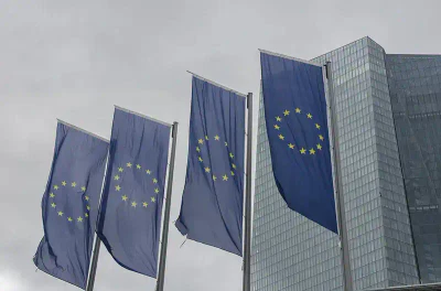 Unter einem bewölkten Himmel wehen vier Flaggen der Europäischen Union vor einem modernen Glasgebäude, vermutlich dem Hauptsitz der EZB. Die Flaggen sind blau und in der Mitte befindet sich ein Kreis aus zwölf goldenen Sternen.