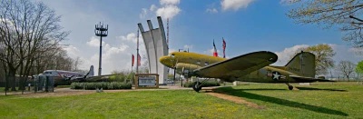 Auf einer grasbewachsenen Außenausstellung sind zwei Militärflugzeuge zu sehen: ein gelbes Flugzeug im Vordergrund und ein silbernes im Hintergrund. Dahinter stehen hohe Gedenksäulen, begleitet von Flaggen und einem Schild, das an den Flughafen Frankfurt erinnert. Links ist ein Kommunikationsturm zu sehen.