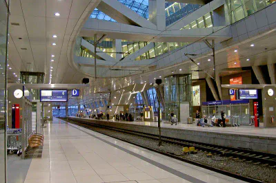 Ein moderner Bahnhof mit elegantem architektonischem Design, mit einem breiten Bahnsteig neben leeren Gleisen, die an den Flughafen Frankfurt erinnern. Auf dem Bahnsteig sind die verschiedenen Bahnsteige und Tore mit den Nummern 6 und D gekennzeichnet. Es gibt Uhren, Bänke und ein paar wartende Leute.
