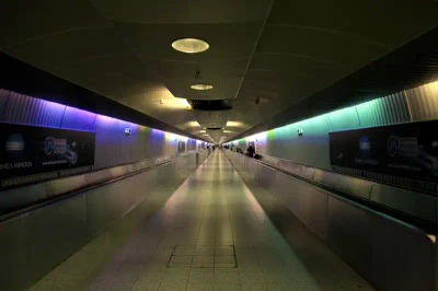 Ein langer, leerer Flughafenkorridor mit einem Laufband, beleuchtet von Decken- und mehrfarbigen Seitenlichtern am Flughafen Frankfurt. An der Decke befinden sich mehrere Scheinwerfer, und an den Wänden hängen Werbeanzeigen. Das Laufband verschwindet in der Ferne und suggeriert Tiefe und Perspektive.