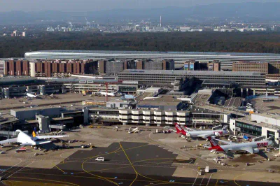Luftaufnahme des geschäftigen Flughafens Frankfurt mit mehreren auf dem Rollfeld geparkten Flugzeugen. Die Terminalgebäude sind groß und modern und haben mehrere Stockwerke. Im Hintergrund sind die Skyline der Stadt und ein Waldgebiet zu sehen. Auf dem Boden sind verschiedene Flughafenfahrzeuge zu sehen.