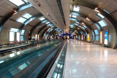 Ein moderner Flughafeninnenraum am Flughafen Frankfurt mit einer langen, gewölbten Decke. Auf der linken Seite verläuft ein Laufband, und mehrere Passagiere sind zu sehen. Große Fenster und Leuchtreklamen säumen die rechte Seite, und der Boden hat eine glänzende, reflektierende Oberfläche.