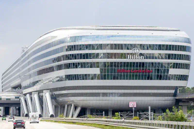 Ein futuristisches, ovales Gebäude mit Außenflächen aus Glas und Metall beherbergt ein Hilton Garden Inn in der Nähe des Flughafens Frankfurt. Das Gebäude wird durch große Säulen erhöht und befindet sich in der Nähe einer befahrenen Straße. Der Hotelname ist deutlich auf der Vorderseite zu sehen.