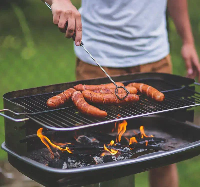 Eine Person grillt Würstchen auf einem Außengrill. Die Person wendet die Würstchen mit einem Grillwerkzeug, während Flammen unter dem Grill sichtbar sind. Die Szene spielt in einem Hinterhof mit grünem Laub im Hintergrund.