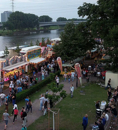 Ein lebhaftes Volksfest am Fluss, bekannt als Höchster Schloßfest, bietet Fahrgeschäfte, Essensstände mit hellen Lichtern und eine Menschenmenge, die das Fest genießt. Im Hintergrund sind Bäume, eine Brücke und einige hohe Gebäude unter einem bewölkten Himmel zu sehen.