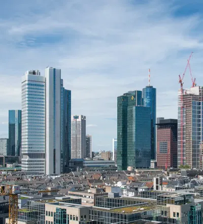 Eine Stadtlandschaft mit modernen Wolkenkratzern unter einem teilweise bewölkten Himmel. Hohe Gebäude mit Glasfenstern dominieren die Skyline, einige davon sind noch im Bau und man sieht Kräne. Im Vordergrund vervollständigen kleinere Gebäude und Dächer die Stadtlandschaft und fangen das Wesen der Innenstadt ein.
