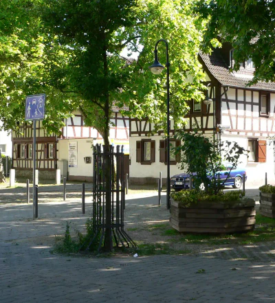 Eine malerische Straßenszene in Unterliederbach besteht aus Fachwerkhäusern mit weißen Wänden und dunklen Holzbalken, üppigen grünen Bäumen und einem blauen Auto, das in der Nähe parkt. Ein hölzerner Blumenkasten mit Grünpflanzen schmückt den Kopfsteinpflasterweg neben einem Laternenpfahl und einem Fußgängerzonenschild.