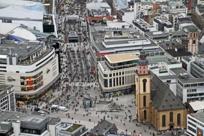 Luftaufnahme eines geschäftigen Stadtgebiets mit dicht gedrängten Gebäuden, Straßen voller Fußgänger und der markanten Kirche mit einem hohen Glockenturm in der Nähe von Zeil. Moderne und historische Architektur verschmelzen und schaffen ein lebendiges Stadtbild voller Aktivität.