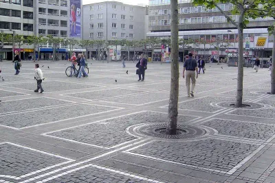 Ein geräumiger städtischer Platz mit ein paar Fußgängern und jemandem, der Fahrrad fährt. Der Platz, der an Zeil erinnert, ist mit grauen Ziegeln gepflastert und mit einem Gitter aus Bäumen bepflanzt. Um den Platz herum stehen mehrere mehrstöckige Gebäude und an einem davon ist ein gut sichtbares Plakat angebracht.