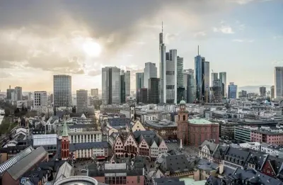 Luftaufnahme von Frankfurt, Deutschland, mit einer Mischung aus historischer und moderner Architektur. Die Skyline zeigt hohe Wolkenkratzer im warmen Abendlicht, darunter fahren Elektroautos durch die Straßen der Stadt, und im Vordergrund stehen historische Gebäude unter einem leicht bewölkten Himmel.