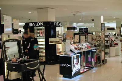 In einer gut beleuchteten Kosmetikabteilung eines Kaufhauses sind Revlon-Produkte, darunter die neue CleanSkin-Linie, prominent vertreten. Auf Regalen und Theken werden verschiedene Make-up-Produkte präsentiert. Ein paar Kunden stöbern in den Artikeln, während ihnen zwei Mitarbeiter helfen.