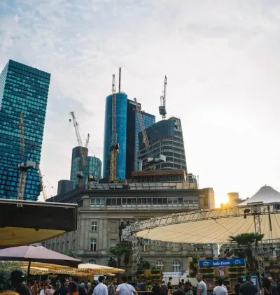 Ein geschäftiger, städtischer Außenbereich mit Menschen, die sich zwischen modernen Wolkenkratzern und Baukränen versammeln. Die Sonne geht unter und taucht die Szene in einen warmen Schein. Sonnenschirme und ein Baldachin spenden Schatten für Tische und Gebäude im Vordergrund und erinnern an die lebhafte Atmosphäre der Freßgass während eines kulinarischen Rundgangs.